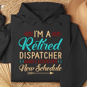 May include: A black hooded sweatshirt featuring the text I'M A RETIRED DISPATCHER AND I LOVE MY NEW SCHEDULE in teal, tan, and red lettering. The design includes small headset icons and horizontal arrow graphics printed on the front chest area.
