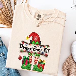 May include: Ivory-colored t-shirt featuring a festive "Dispatcher Elf" design. The graphic includes an elf hat, string lights, and gift boxes. The text "Dispatcher Elf" is prominently displayed. The shirt has a crew neck and short sleeves.