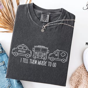May include: A dark gray t-shirt with a white outline of a police car, fire truck, and ambulance. The text "I tell them where to go" is printed below the vehicles.