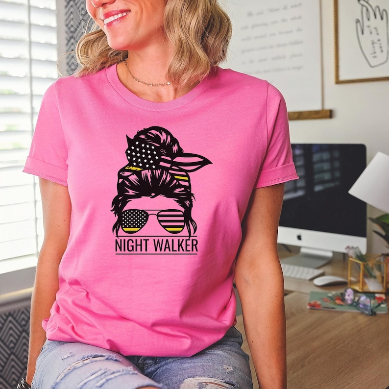 May include: Pink t-shirt featuring a black and yellow graphic of a woman with a messy bun, sunglasses, and a bandana. The bandana is patterned with the American flag. The text "NIGHT WALKER" is below the graphic.