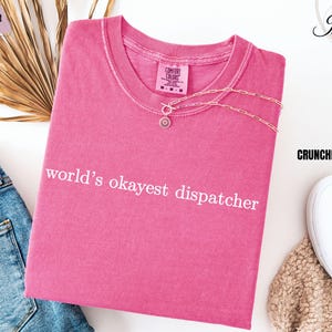 May include: Pink t-shirt with the text "world's okayest dispatcher" in white lettering.