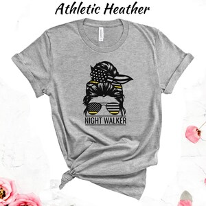 May include: Grey t-shirt featuring a graphic of a woman with a messy bun, sunglasses, and a bandana. The bandana and sunglasses have a yellow and black stripe pattern. The text "NIGHT WALKER" is below the graphic.