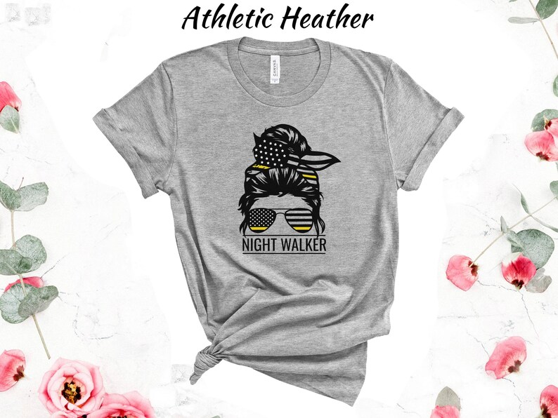 May include: Grey t-shirt featuring a graphic of a woman with a messy bun, sunglasses, and a bandana. The bandana and sunglasses have a yellow and black stripe pattern. The text "NIGHT WALKER" is below the graphic.