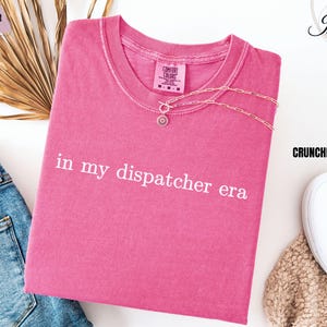 May include: Pink t-shirt with the text "in my dispatcher era" in white lettering.