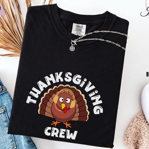 May include: Black t-shirt with a Thanksgiving-themed graphic. The design features a cartoon turkey wearing headphones, surrounded by the words "THANKSGIVING CREW" in white. A silver necklace is draped across the shirt.