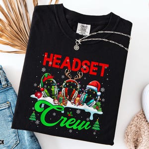May include: Black t-shirt with the words "HEADSET Crew" in red and green, featuring headset designs decorated with Christmas elements. A silver necklace is draped across the shirt, and a pair of blue jeans is visible.