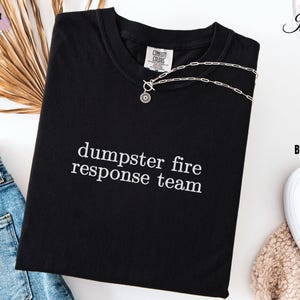 May include: Black t-shirt with the text "dumpster fire response team" in white. The shirt has a crew neck and short sleeves. A silver necklace with a pendant is draped across the shirt. The shirt is folded on a white surface.