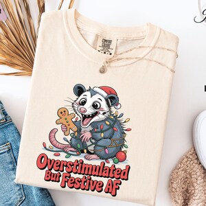 May include: Ivory-colored t-shirt featuring a cartoon opossum wearing a Santa hat and wrapped in Christmas lights, holding a gingerbread man. The text "Overstimulated But Festive AF" is printed in red and white.