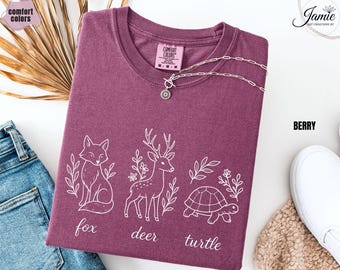 May include: Berry-colored t-shirt with white line art of a fox, deer, and turtle, with the words "fox", "deer", and "turtle" below each animal. The shirt has a crew neck and is folded. A silver necklace with a pendant is on top of the shirt.