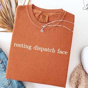 May include: A rust-colored t-shirt with the text "resting dispatch face" printed in white on the front.