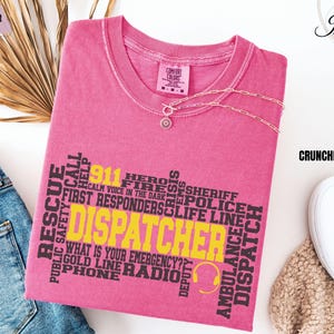 May include: A pink t-shirt with the word "DISPATCHER" in yellow, surrounded by related words like "911", "RESCUE", and "AMBULANCE". The shirt also features a headset graphic. The shirt is part of the "Comfort Colors" collection.