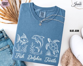 May include: Blue jean colored t-shirt with white illustrations of a fish, dolphin, and turtle. The words "Fish Dolphin Turtle" are printed below the images. A silver necklace is draped across the shirt.
