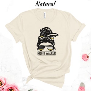 May include: A beige t-shirt featuring a black and yellow graphic of a woman with a messy bun, sunglasses, and the text "NIGHT WALKER".