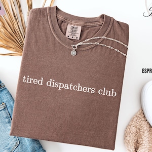 May include: A brown t-shirt with the text "tired dispatchers club" printed in white. The shirt is laid flat on a white surface with a pair of white sneakers and a brown fuzzy blanket in the background.