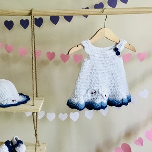 May include: White crocheted baby dress with blue trim and a blue bow on the bodice. The dress is hanging on a wooden hanger with a pink and white heart garland in the background.
