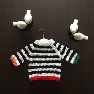 May include: A striped crocheted baby sweater with orange, green, and red trim. The sweater is hanging on a hanger.