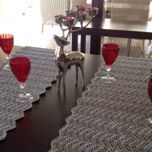 Puede incluir: Dos caminos de mesa de crochet grises con bordes festoneados sobre una mesa de madera oscura. Cuatro copas de vino tinto están sobre los caminos de mesa. Un candelero de plata en forma de reno con velas encendidas está en el centro de la mesa.