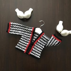 May include: A black and white crocheted cardigan with red trim. The cardigan has a button closure and is hanging on a hanger.