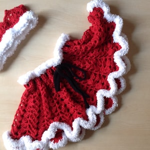 May include: A red and white crocheted Christmas outfit for a baby, featuring a skirt with a white trim and a matching headband.