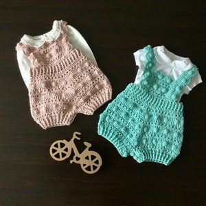 May include: Two handmade crochet baby rompers in pink and teal, each paired with a white shirt. The pink romper has a lace collar, while the teal one has a scalloped collar. A wooden bicycle decoration is also present.