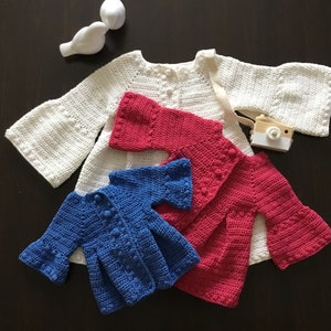May include: Three crocheted baby cardigans in white, pink, and blue. The cardigans have bell sleeves and button closures.
