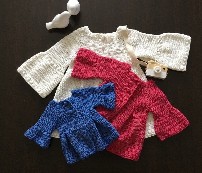 May include: Three crocheted baby cardigans in white, pink, and blue. The cardigans have bell sleeves and button closures.