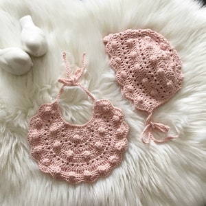 May include: A pink crocheted baby bonnet and bib set with a textured, raised dot pattern.
