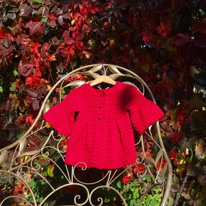 May include: A red crocheted cardigan with bell sleeves, hanging on a metal chair. The cardigan has a button closure and is made with a soft, textured yarn.