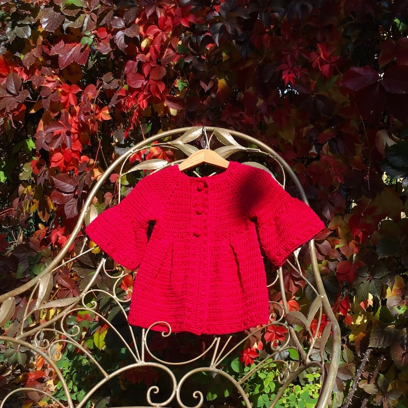 May include: A red crocheted cardigan with bell sleeves, hanging on a metal chair. The cardigan has a button closure and is made with a soft, textured yarn.