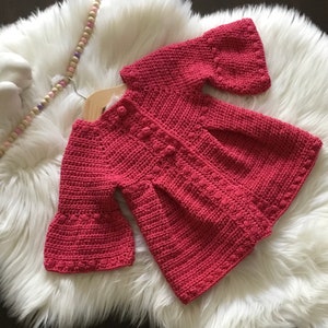 May include: A crocheted baby cardigan in a dusty rose color with bell sleeves and a button closure.