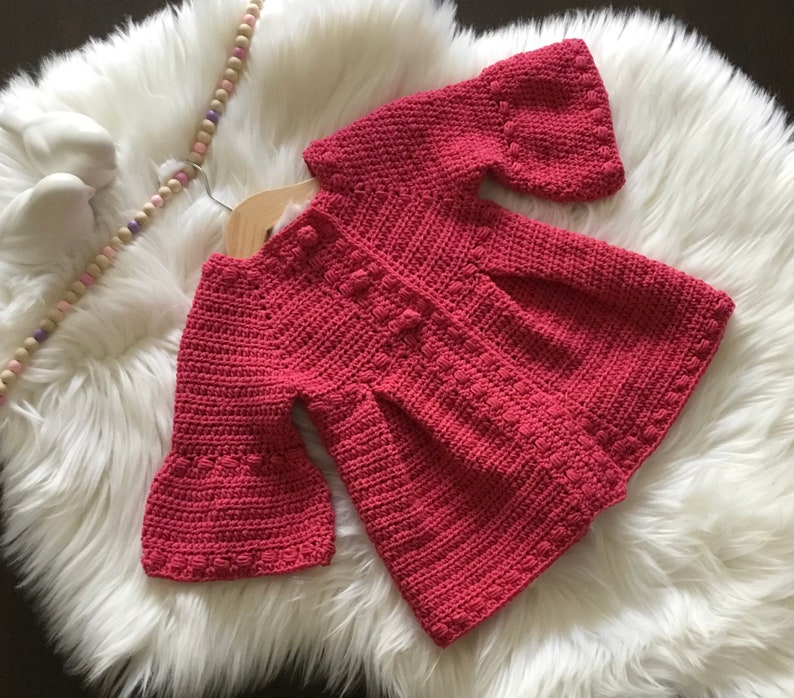 May include: A crocheted baby cardigan in a dusty rose color with bell sleeves and a button closure.