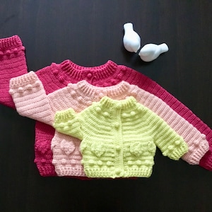May include: Three crocheted cardigans in pink, light pink, and lime green with heart-shaped details and button closures.