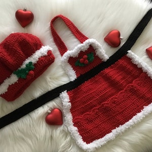 May include: A red crocheted Santa hat and apron set with white trim and green holly accents. The set includes a black strap for the apron.