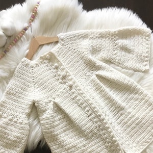 May include: A white crocheted cardigan with bell sleeves. The cardigan has a button closure and a delicate stitch pattern.
