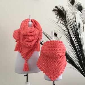 Może przedstawiać: Dwa trójkątne szale wykonane na szydełku z frędzlami, jeden większy od drugiego, oba w kolorze koralowego różu. Szale są prezentowane na białych manekinach na białym tle.
