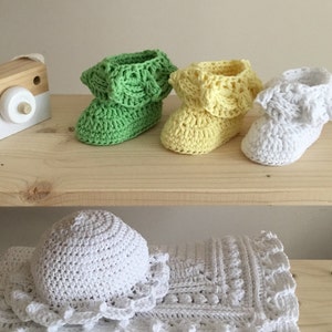 May include: Three pairs of crocheted baby booties in green, yellow, and white. The booties have a ruffled edge and are displayed on a wooden shelf.