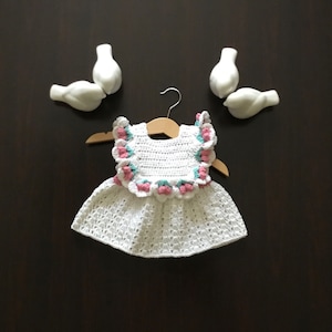 Puede incluir: Vestido de bebé de crochet blanco con ribete floral rosa y verde. El vestido está colgado en una percha de madera.
