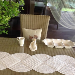 May include: A white crocheted table runner with a wavy design sits on a brown wicker table with a white ceramic serving dish and a white coffee cup.