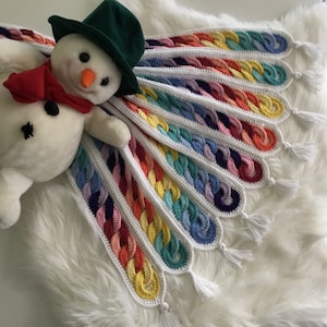 Könnte beinhalten: Eine Regenbogen-Häkeldecke mit weißen Rändern und Quasten. Die Decke ist mit einem Kettenmaschenstichmuster gearbeitet und hat einen gewellten Rand.