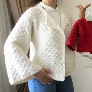 May include: A white crocheted cardigan with a shawl collar and long sleeves. The cardigan is open at the front and has a loose fit. The cardigan is being held by a person wearing blue jeans.
