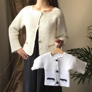 Puede incluir: Dos cárdigans de punto blanco, uno más grande y otro más pequeño. El cárdigan más grande tiene un cierre de botón dorado y un bolsillo a cada lado. El cárdigan más pequeño tiene botones azules y ribetes azules en los bolsillos.