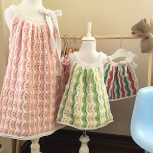 Puede incluir: Tres vestidos de crochet en rosa, verde y rojo con rayas blancas. Los vestidos están colgados en un estante de madera.