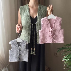 Może przedstawiać: Trzy dzianinowe kamizelki w różnych kolorach: zielonym, różowym i szarym. Kamizelki są bez rękawów i mają dekolt w kształcie litery V. Zielona kamizelka ma zapięcie na sznurek przy szyi. Różowa i szara kamizelka mają zapięcie na guziki z przodu.