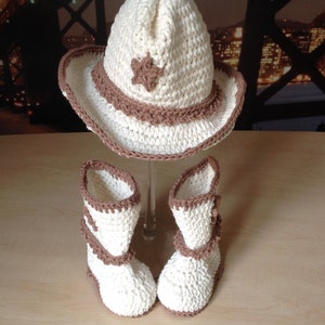 May include: A crocheted cowboy hat and booties set in white and brown. The hat has a star design and the booties have a brown trim.