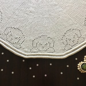 May include: White crocheted doily with a scalloped edge. The doily is on a dark brown surface with small white snowflakes scattered around it.