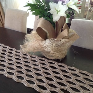 Puede incluir: Un camino de mesa de crochet beige con un patrón de diamante sobre una mesa de madera marrón oscuro. Lirios blancos en un jarrón de papel marrón con fibra de sisal están sobre la mesa.