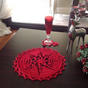 May include: Red crocheted doily on a dark wood table with a red wine glass and a silver reindeer figurine.