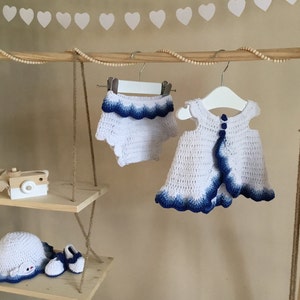 May include: A white crocheted baby dress and matching diaper cover with blue ombre trim. The set is hanging on a wooden rack with a white heart garland above.