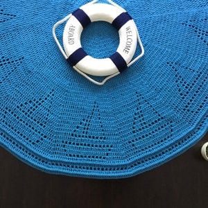 May include: A blue crocheted round rug with a white life preserver with the words "Welcome" and "Aboard" printed on it. The rug is on a dark brown surface.