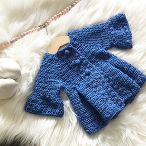 May include: A blue crocheted baby cardigan with a button closure and ruffled sleeves. The cardigan is laid out on a white fluffy surface.